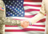 USA military man in uniform and civil man in suit shaking hands with adequate national flag on background United States of America