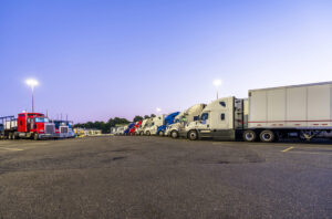 Different big rig semi trucks with semi trailers standing in row on truck stop parking lot at evening time for rest of drivers and safety requirements for freight forwarders