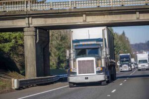 Big rig classic semi truck with covered semi trailer driving on the road under the bridge in front of semi trucks convoy