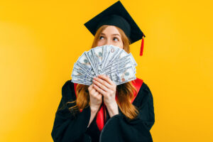 beautiful graduate girl, in a black gown, and a black master's hat, with dollars in her hands, marks the graduation, on a yellow background