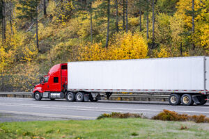 Red long hauler big rig semi truck transporting cargo in dry van semi trailer driving on the autumn road with yellow trees on the hill