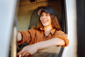 A woman with brown hair and a cap beams with joy while sitting in the driver's seat of a large truck.