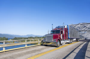 Burgundy big rig classic semi truck with refrigerated shiny semi trailer transporting commercial cargo on the Bridge of God in Columbia River Gorge