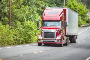 Red big rig semi truck with refrigerator semi trailer climbing uphill on the winding forest road with green trees