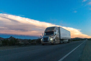 At sunset, a mighty semi truck rumbled through the Nevada desert, its massive form cutting through the vast landscape. The mountains stood stoically in the background, adding a touch of grandeur to the scene.