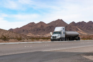 Against the backdrop of a breathtaking sunset, a white semi truck traversed the vast Nevada Desert, its silhouette cutting through the landscape, with majestic mountains rising in the distance.