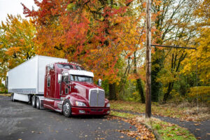 Burgundy classic big rig semi truck tractor with high cab with roof windows and dry van semi trailer standing on the city street with autumn red and yellow maple trees