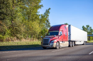 Red big rig commercial semi truck transporting cargo in refrigerator semi trailer running on the wide road in sunny day