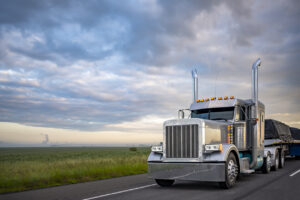 Beautiful classic big rig American semi truck with light transporting covered cargo on flat bed semi trailer driving on the twilight highway road with stormy clouds sky