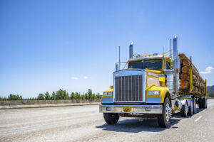 Bright yellow shiny day cab bonnet big rig semi truck transporting loaded with logs semi trailer driving on the wide multiline highway road