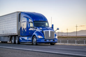 Bright blue powerful big rig semi truck transporting cargo in reefer semi trailer driving on the road with sunset