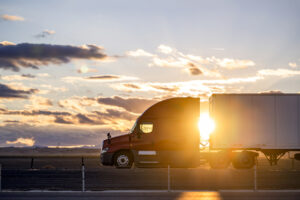 Professional bonnet red big rig semi truck transporting cargo in dry van semi trailer driving on the road on the California plato at sunset time