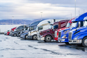 Big rigs semi trucks with semi trailers standing in row on truck stop parking lot with wet surface waiting for the continuation freights