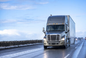 Gray big rig semi truck with turned on headlights transporting cargo in dry van semi trailer running on the wet evening road with rain and reflection on the road surface