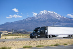 Stylish black big rig bonnet semi truck tractor transporting cargo in refrigerator semi trailer running on the highway road past the snowy mountain in California