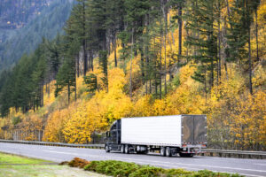 Black long haul big rig semi truck transporting cargo in refrigerator semi trailer driving on the picturesque highway road with autumn yellow mountain hill in Columbia Gorge