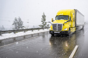 Bright yellow big rig semi truck transporting cargo in dry van semi trailer driving on the wet slippery highway road with winter snow storm