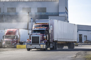 Classic burgundy big rig semi truck tractor with chrome parts and container on semi trailer running on the road in industrial area raising a cloud of dust