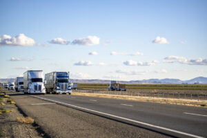 Convoy of the industrial big rig semi trucks with semi trailers transporting cargo running on the divided highway road in California