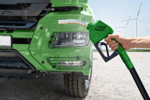 Male hand is holding biofuel filling nozzle in the background of green truck. The truck parked in the background of wind turbines.