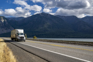 Day cab white big rig semi truck with huge roof spoiler transporting cargo in dry van semi trailer running on the highway road along the Columbia River with mountains on the sides