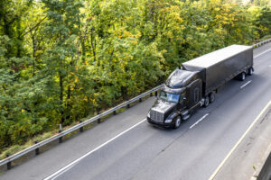 Stylish black big rig semi truck with extended cab transporting cargo in conestoga flat bed railer running on the autumn highway road with forest on the side