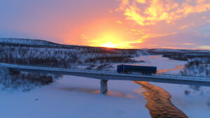 AERIAL: Semi truck crossing the bridge above icy river in the winter at sunset