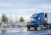 Big rig blue semi truck tractor transporting commercial cargo in refrigerator semi trailer going on the wet road with melting snow with winter snowy trees on the side