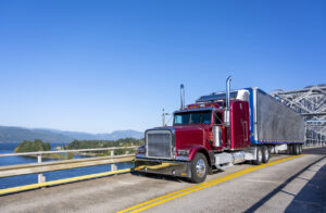 Red classic bonnet big rig semi truck transporting frozen cargo in bright refrigerated semi trailer driving on the truss bridge across the river