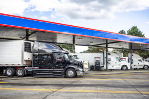 Stylish black big rig semi truck with refrigerated semi trailer standing with another semi trucks on the truck stop fuel station for refile the truck tanks