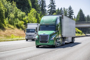 Green big rig semi truck with dry van semi trailer and small rig cab over semi truck with box trailer running side by side on the multiline highway road