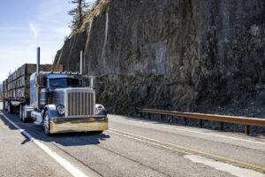 Classic stylish black big rig semi truck tractor transporting boxes for carry vegetables and fruits on flat bed semi trailer running on the mountain narrow road