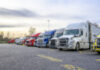 Different big rigs semi trucks with semi trailers standing in row on truck stop parking lot with reserved spots for truck driver rest and compliance with established truck driving regulations