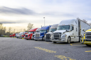 Different big rigs semi trucks with semi trailers standing in row on truck stop parking lot with reserved spots for truck driver rest and compliance with established truck driving regulations