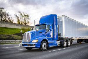 Powerful blue day cab big rig semi truck with dry van semi trailer driving on the wide road at twilight time