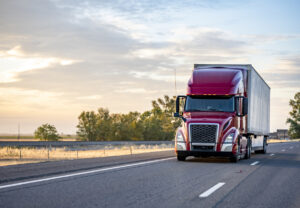 Popular model burgundy big rig semi truck transporting commercial cargo in dry van semi trailer running on the evening highway road with sunshine