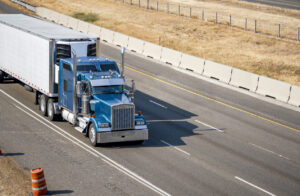 Classic idol blue big rig semi truck with chrome parts and cab with truck driver rest compartment with upper windows transporting cargo in refrigerated semi trailer running on the wide highway road