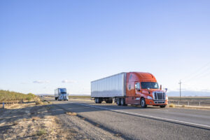 Team of two orange and blue big rigs semi trucks transporting cargo in dry van semi trailers running together on the flat straight highway road in California