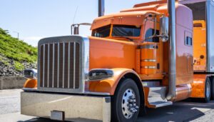 Bright orange big rig classic American bonnet semi truck tractor with painted accessories transporting cargo in stainless steel dry van semi trailer driving on the road at sunny day