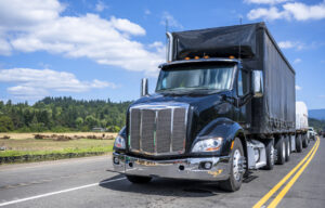 Black day cab big rig semi truck transporting cargo on dry van and flat bed semi trailers driving on the narrow road at sunny day