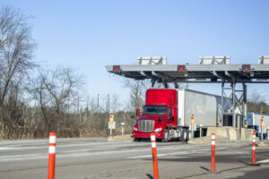 Red day cab big rig semi truck with dry van semi trailer passes a toll booth on a toll highway road