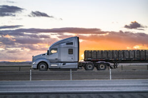 Profile of the gray big rig semi truck with extended cab transporting boxes on the flat bed semi trailer driving on the flat highway road with sunset sky