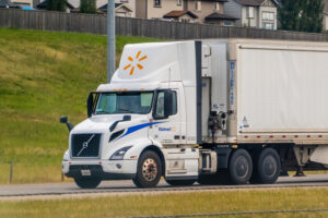 A white Volvo semi truck with a Walmart trailer captured in motion on a highway, with suburban homes visible in the bg.