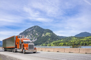 Stylish classic orange big rig semi truck bonnet tractor with same color reefer semi trailer trim and refrigerator unit transporting cargo running on the road along the river