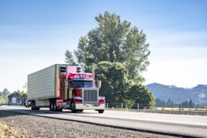 Popular classic bonnet American idol big rig red semi truck with extended cab transporting cargo in refrigerated semi trailer driving on the flat highway road at sunny day