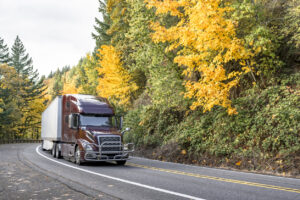 Shiny brown big rig semi truck with dry van semi trailer driving on the winding narrow road with autumn trees on the hillsides