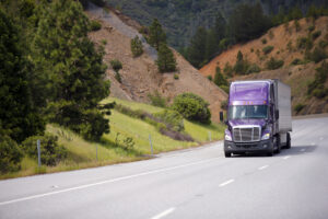 Lilac semi truck with aluminum trailer is moving along winding highway through the pass in California against the background of orange mountain slopes