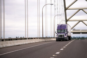 Lilac modern big rig semi truck with trailer moves along the arch bridge to deliver commercial cargo